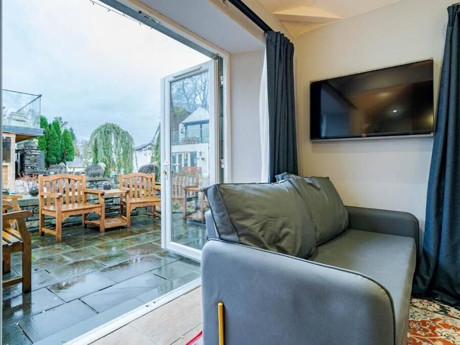 A modern living room with a grey sofa, a flat-screen TV mounted on the wall, and a sliding glass door leading to an outdoor patio with wooden furniture.