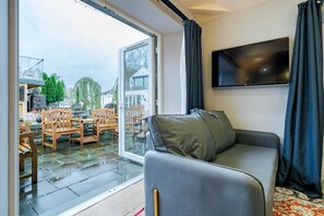 Ett modernt vardagsrum med en grå soffa, en platt-TV som är monterad på väggen och en skjutdörr i glas som leder ut till en uteplats med trädgårdsmöbler.