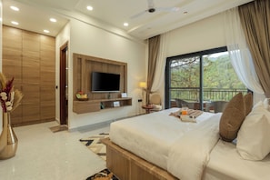 A bedroom with a large bed, a TV mounted on the wall, a balcony with chairs, and a wooden wardrobe.