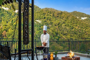A chef cooking outdoors with a view of a forested hillside. - ELIVAAS Panchvati Veloraa 3BHK (Solan)