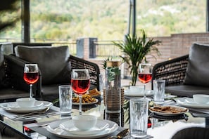 Une table à manger dressée avec des assiettes, des bols, des verres et des couverts, donnant sur un paysage verdoyant par la fenêtre.