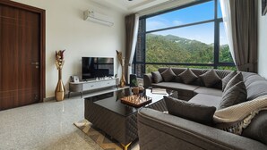 A modern living room with a large window, a flat-screen TV, a sofa, and a coffee table. - ELIVAAS Panchvati Veloraa 3BHK (Solan)
