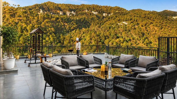 A terrace with wicker furniture, a glass table, and a view of a forested hillside. - ELIVAAS Panchvati Veloraa 3BHK (Solan)