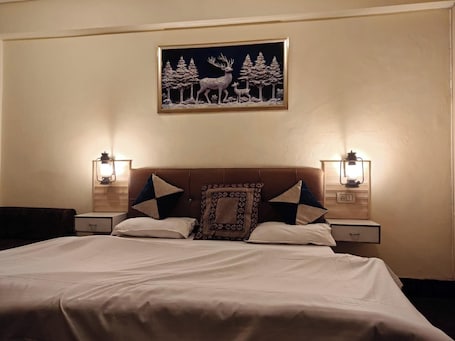 A neatly made bed with a patterned pillow, two bedside tables with lamps, and a framed picture of deer and trees on the wall.. Travel Meerut Inn
