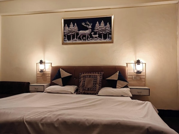 Una cama bien hecha con un cojín estampado, dos mesitas de noche con lámparas y un cuadro en la pared con ciervos y árboles.