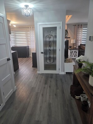 Un pasillo con una puerta que indica 'PANTRY', piso de madera, una lámpara en el techo y un rincón de estar con un sofá y una mesita.