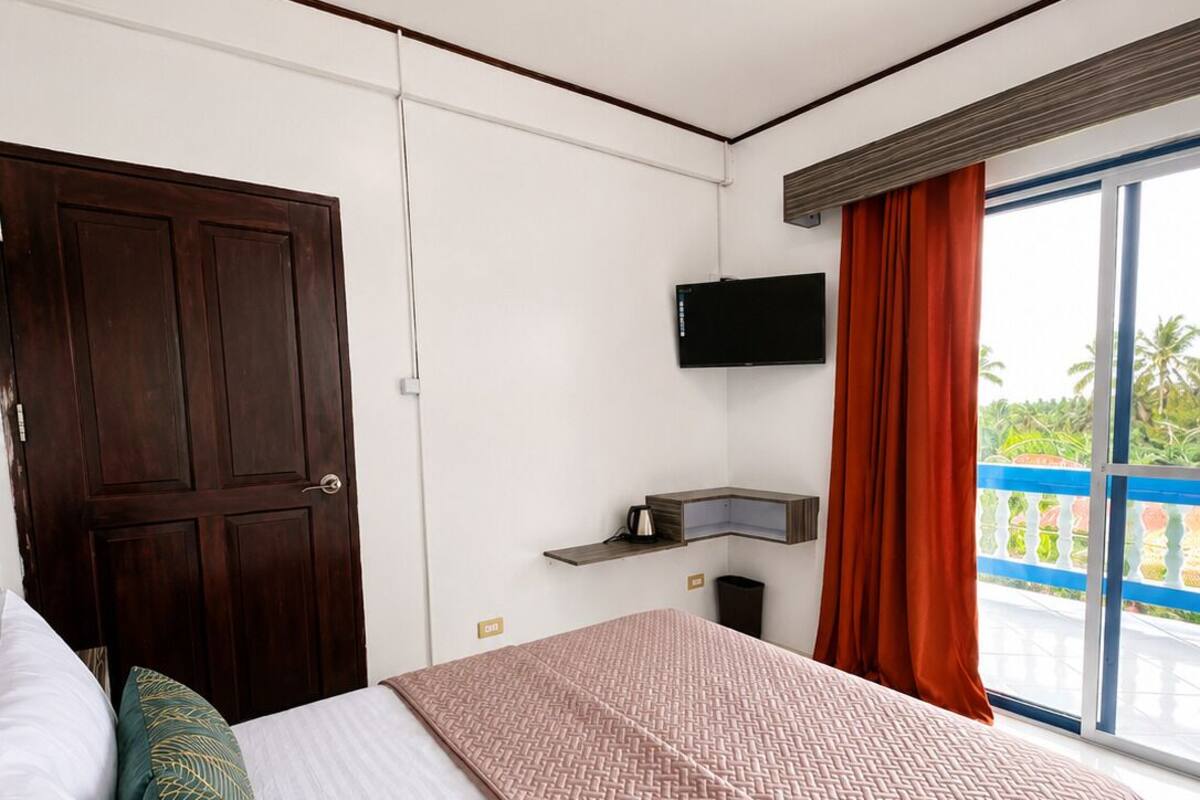 A bedroom with a bed, a TV, a window with curtains, a door, and a small shelf with a kettle.