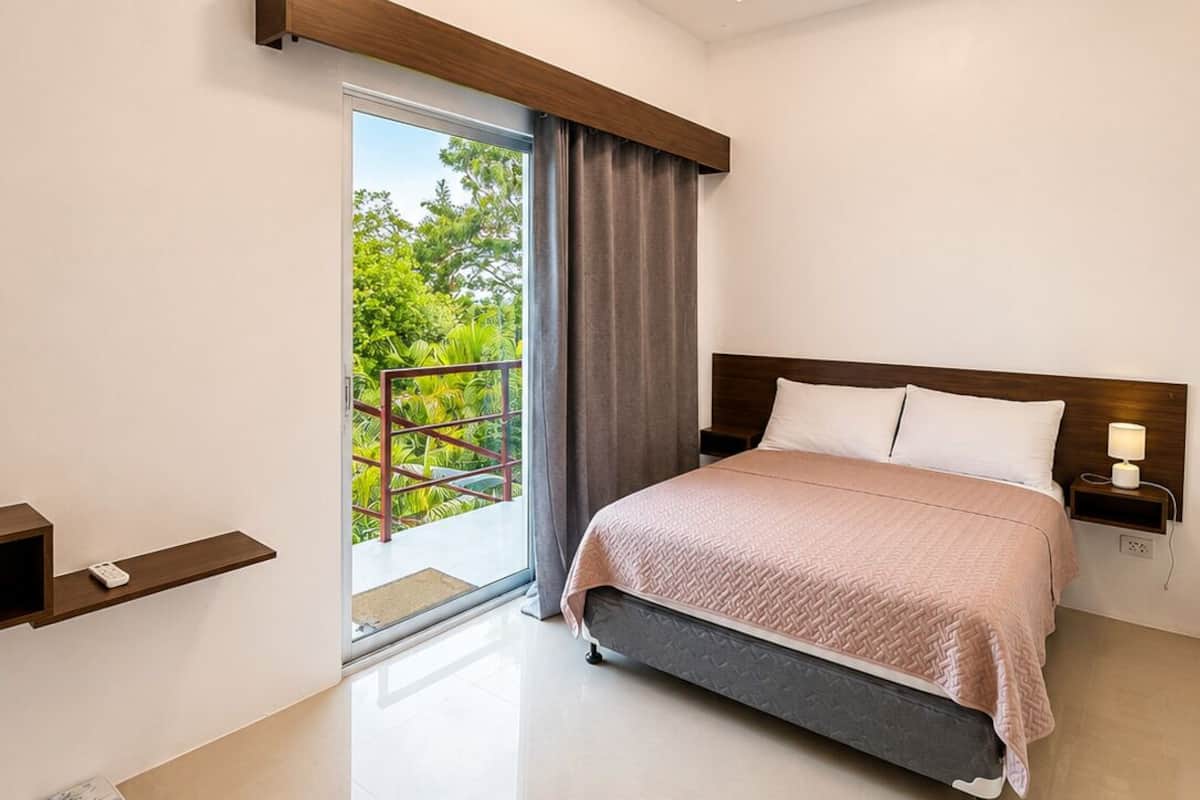 A modern bedroom with a bed, bedside table, and a view of greenery outside.