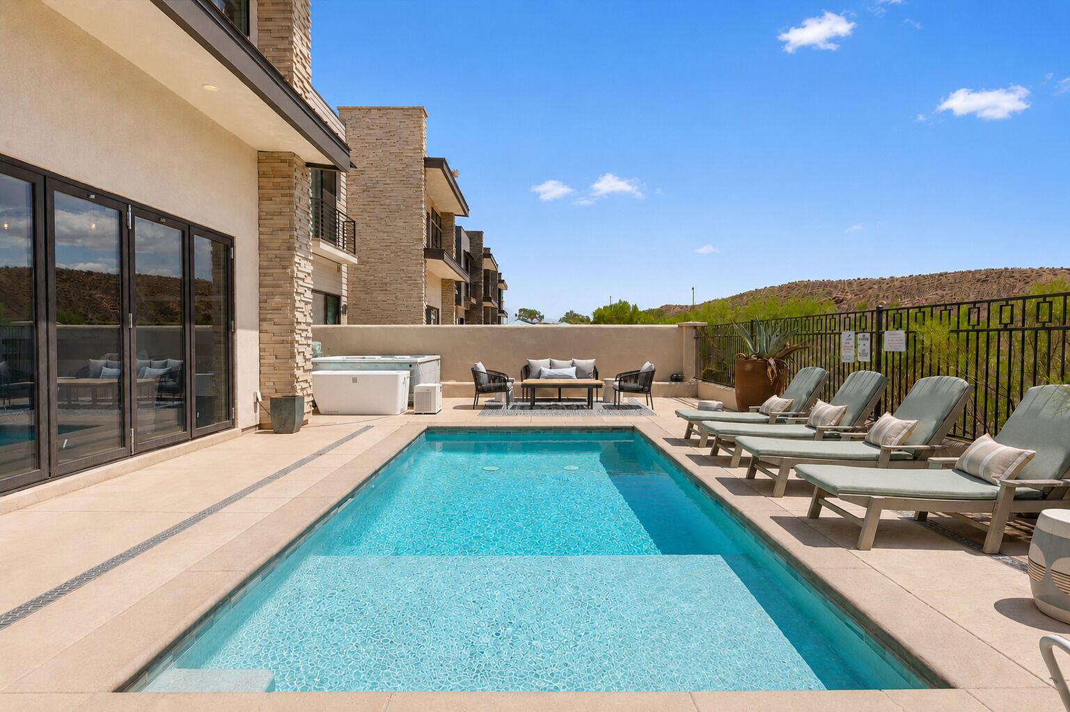 Une piscine rectangulaire, agrémentée de chaises longues et d’une table, est entourée d’un bâtiment doté de grandes fenêtres et d’une façade en pierre.