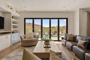 A modern living room with a sofa, coffee table, and TV, large sliding glass doors to a pool area, and a kitchen with white cabinets and a TV. - St. George Oasis 7 Bedroom Home (Washington)