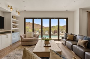 Una sala de estar moderna con sofá, mesa de centro y televisión, puertas corredizas de vidrio que dan a una zona de piscina, y una cocina con armarios blancos y televisión.