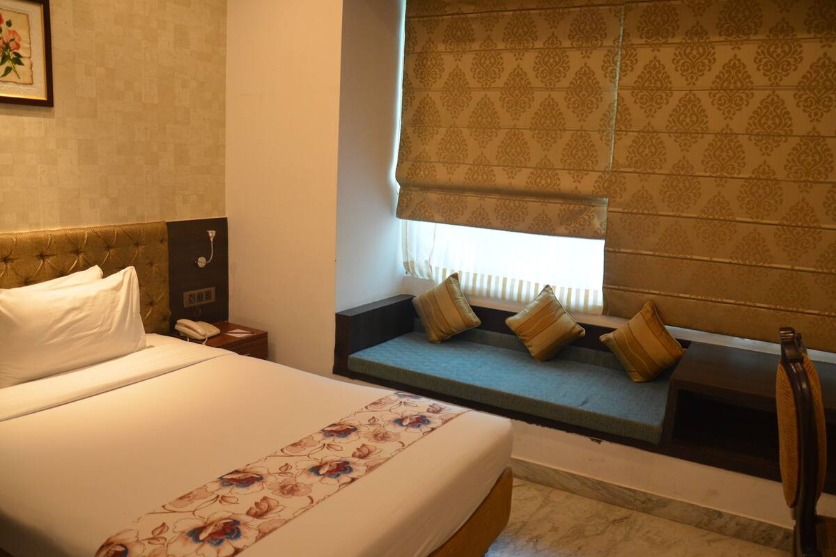 A hotel room with a bed, bedside table, lamp, window with blinds, and a built-in bench with cushions.