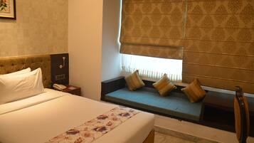 Habitación de hotel con cama, mesita de noche, lámpara, ventana con persianas y un banco empotrado con cojines.