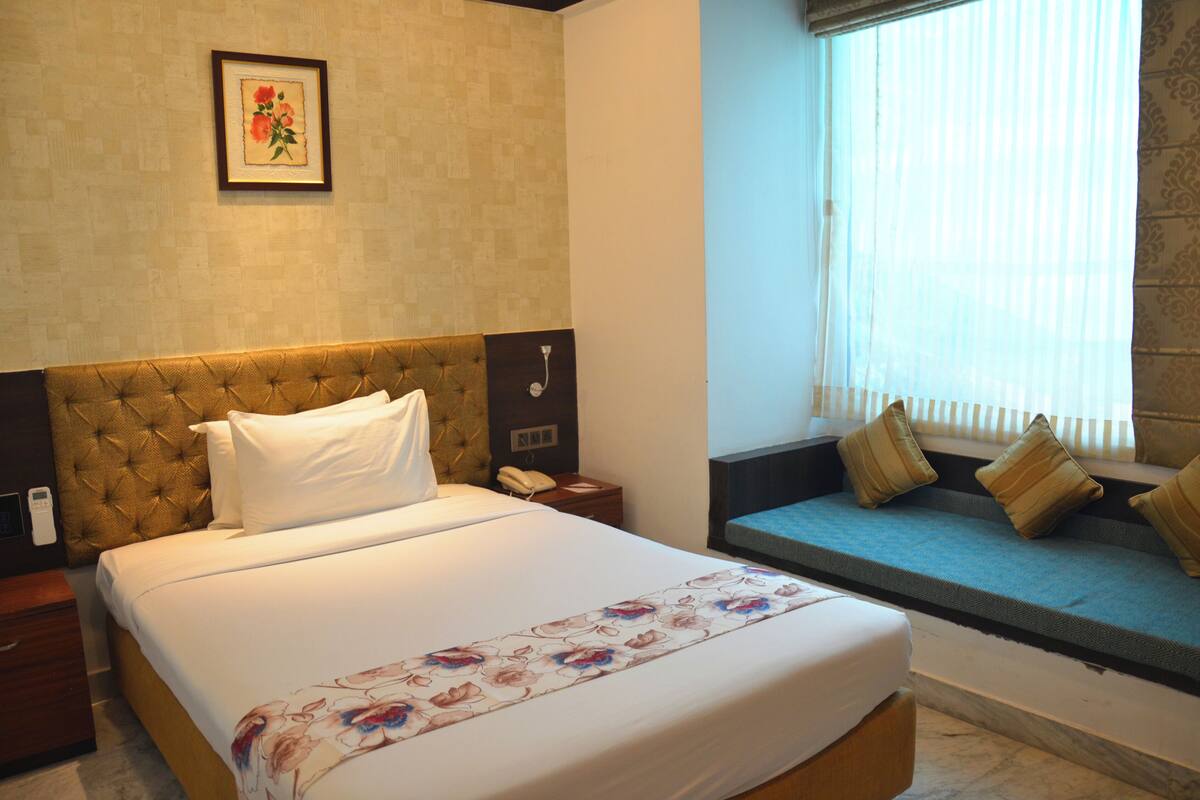 A hotel room with a large bed, a bedside table, a nightstand, a window with blinds, and a wall-mounted telephone.