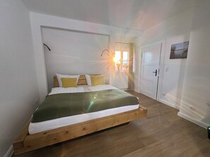 Ein Schlafzimmer mit einem hölzernen Bett, weißer Bettwäsche und einem Fenster, durch das Sonnenschein fällt.