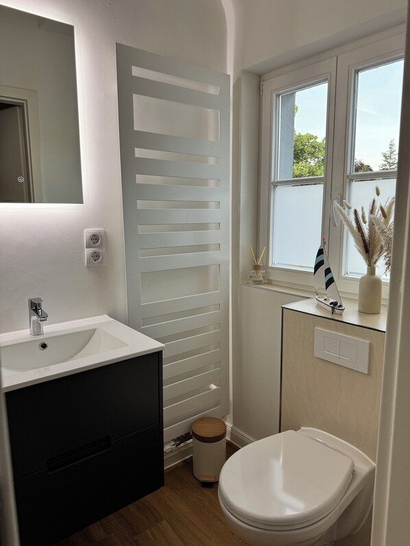 Ein Badezimmer mit einem weißen Waschbecken, einem Spiegel und einer Toilette. Ein Fenster bietet Blick auf Grünflächen.