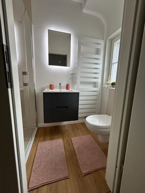 Ein Badezimmer mit einem weißen Waschbecken, einem dunklen Schrank und einer weißen Toilette. Auf dem Boden liegen zwei rosa Fußmatten.