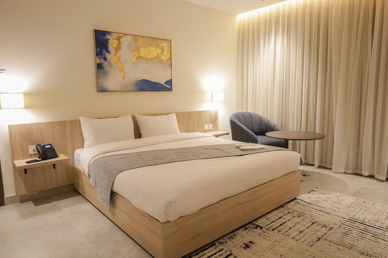 A hotel room with a large bed, a chair, a small table, and a painting on the wall.