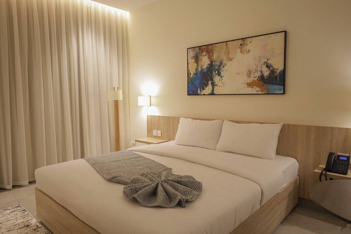 Quarto de hotel com cama, luminária, telefone, quadro na parede e janela com cortinas.