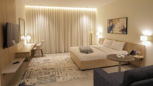 A hotel room with a bed, bedside tables, a desk, a chair, a TV, a painting, and a small round table. - ISHQ Hotel (Aqaba)