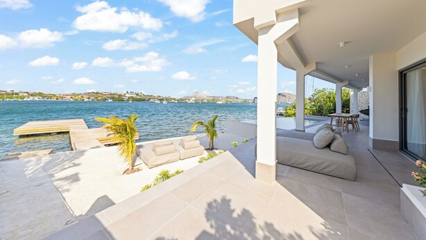 Een modern terras buiten, met uitzicht op zee, een houten steiger en een zithoek met kussens.