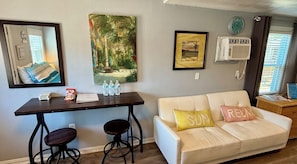 Ein Wohnzimmer mit einem weißen Sofa, zwei Barhocker, einem Holztisch mit Wasserflaschen, Snacks und gerahmten Bildern an der Wand.