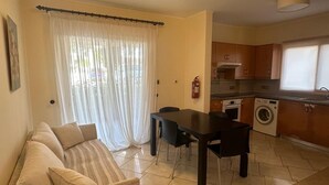 A living room with a sofa, a dining table, and a kitchen area with a washing machine. - Elysia Park Pafos (Paphos)
