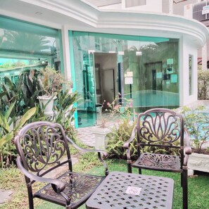 A courtyard with two ornate metal chairs and a small table, surrounded by potted plants and a glass building entrance. - 2-bedroom condo in brilliant Muang Pattaya with AC and gym access  Stream room (Muang Pattaya)
