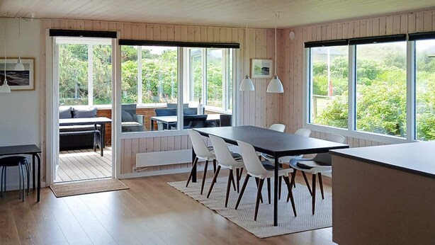 Una cocina moderna con suelo de madera, una zona de comedor con sillas blancas y ventanas amplias que ofrecen vistas a zonas verdes.