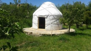 A white yurt with a wooden door, surrounded by a wooden deck, set in a grassy area with trees. - Belzhan Yurt Lodge (Grigorevka)