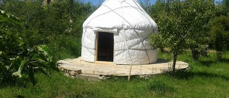 A white yurt with a wooden door, surrounded by a wooden deck, set in a grassy area with trees.