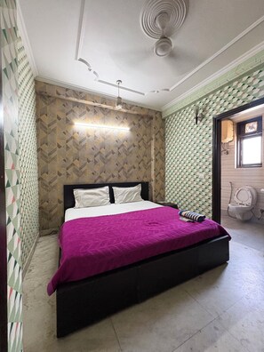 Une chambre Ă coucher avec un lit, un ventilateur de plafond, un papier peint Ă motifs et une fenĂȘtre donnant sur des toilettes.