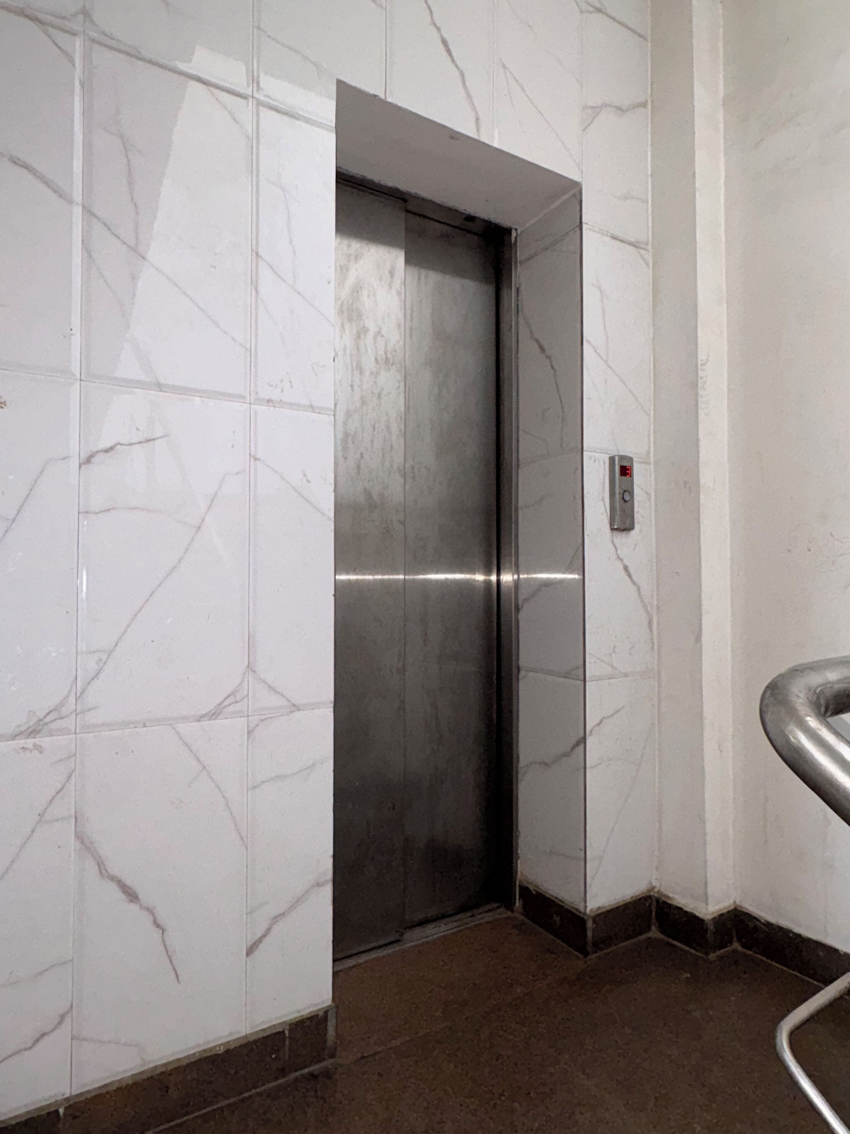 An elevator in a hallway with white marble walls and a brown floor.