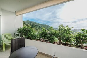 A balcony with a table and chairs, a view of a green landscape with trees and hills, and a clear sky. - Panoramic 1BR, Pool, Sauna & Sea Views C196 (Karon)