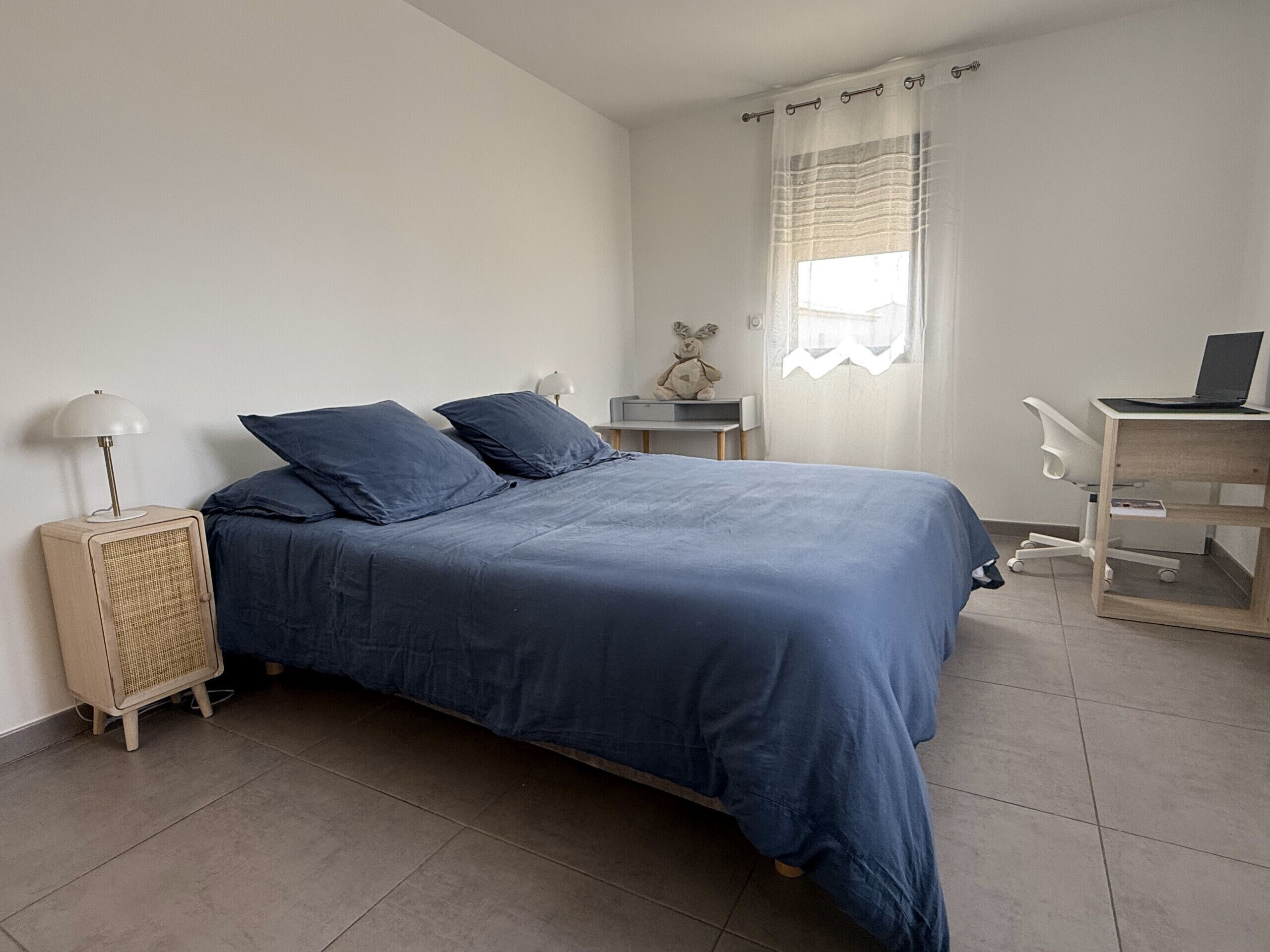 Een slaapkamer met een bed, een bureau met een laptop, een lamp en een klein tafeltje met een teddybeer.