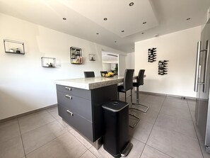 Eine moderne Küche mit einer zentralen Kochinsel, schwarzen Stühlen, einem Kühlschrank und Weinflaschen an der Wand.