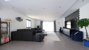 A modern living room with a sectional sofa, a coffee table, a bookshelf, a potted plant, and a kitchen area with cabinets and a countertop. - 12-Person Villa A/C Heated Pool office close Aix Provence (Salon-de-Provence)