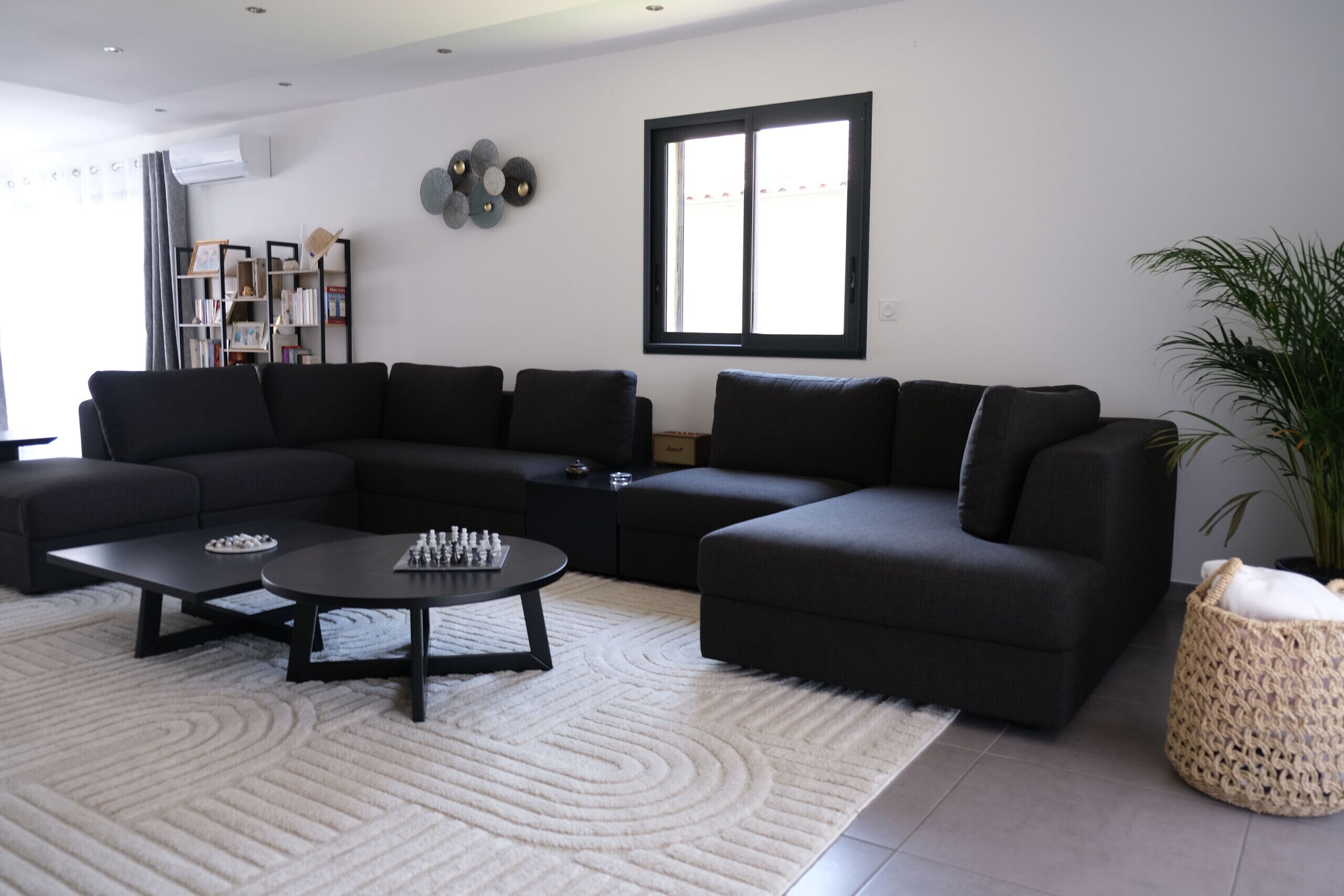 A modern living room with a sectional sofa, a coffee table, a bookshelf, and a large window.