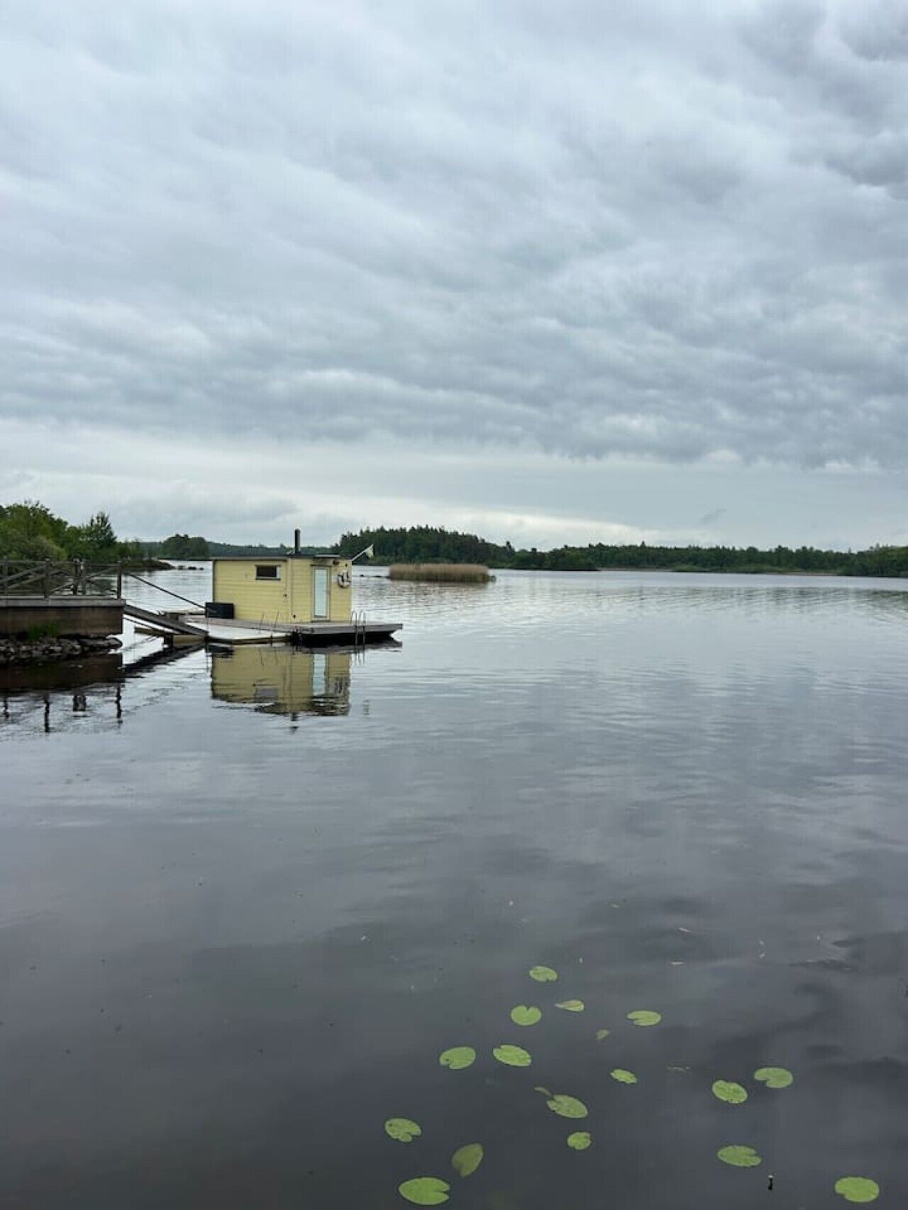 A calm lake with a dock and a small building, surrounded by trees and overcast sky.