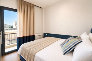 A hotel room with a bed, a view of the city through a balcony, and a window with curtains. - LIV - Upscale Athenian Apartment 4C (Athens)