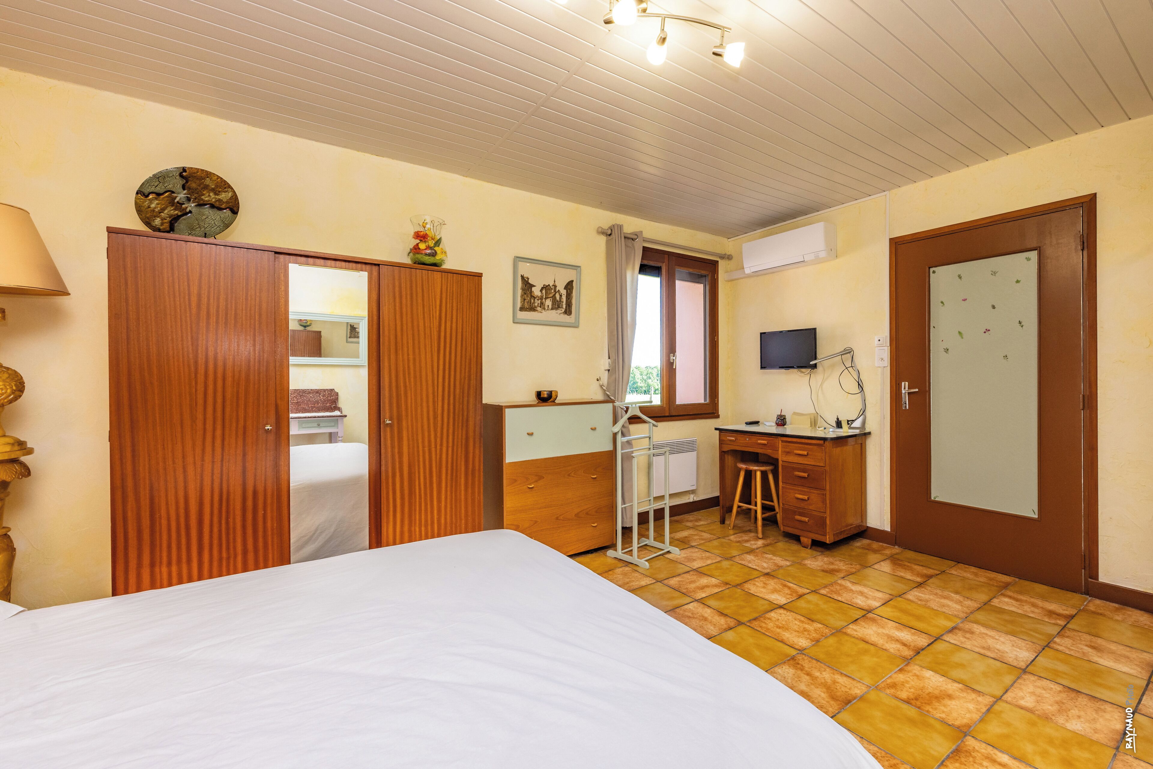 A bedroom with a bed, wooden wardrobe, desk, chair, and a window with curtains.