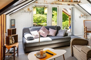 Ein Loft-Wohnzimmer mit grauem Sofa, Korbstühlen, einem kleinen Tisch und Blick auf Grün durch große Fenster.