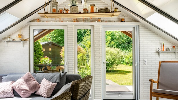 Una habitación acogedora con techo inclinado, un sofá con cojines, un sillón de mimbre y vistas a un jardín por una puerta de cristal.