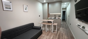 Een moderne woonruimte met een bank, een eettafel met krukjes, een keuken met betegelde spatwand en een wandgemonteerde televisie.