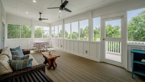 Eine geräumige, überdachte Veranda mit Holzboden, weißen Wänden und großen Fenstern mit Blick auf viel Grün. Es gibt ein Rattangeflechtsofa mit Kissen, einen kleinen Holztisch und einen Deckenventilator.