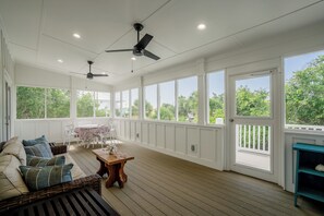 A spacious covered porch with a wooden floor, white walls, and large windows offering a view of greenery. There is a wicker sofa with cushions, a small wooden table, and a ceiling fan. - About Time-3BR/4BA Pet Friendly,Private Pool (St. George Island)