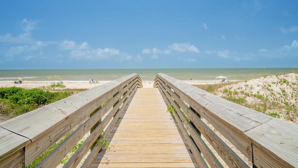 Ein hölzerner Steg führt zu einem Strand bei blauem Himmel.