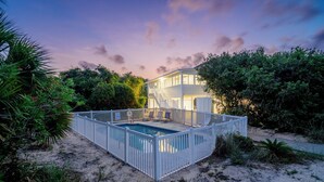 Ein modernes Haus mit Pool, umgeben von einem weißen Zaun, in einer sandigen Umgebung mit üppiger Vegetation.