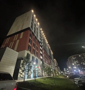 Un immeuble de grande hauteur, doté de nombreux étages, de fenêtres illuminées et d’un parking où sont garées des voitures.