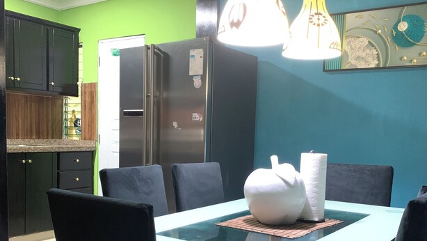 Un'area pranzo con un tavolo in vetro, sedie nere, un vaso bianco e un frigorifero sullo sfondo.